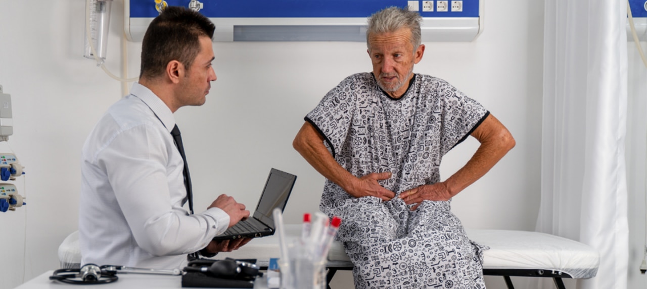 Elderly male patient sits on exam table speaking to doctor about hernia pain