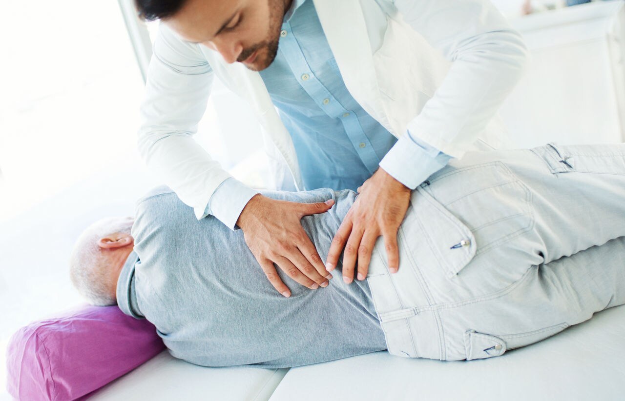 Closeup side view of an early 60's senior gentleman having some lower back pain. He's at laying sideways on the examination table while the doctor is touching and massaging his lower back area.